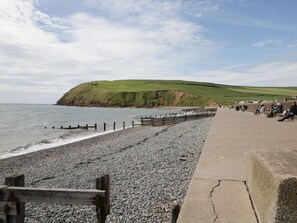 A pebbled beach with a wooden walkway and a grassy hill in the background. - Holly Cottage (St Bees)