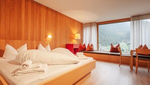 Ein modernes Hotelzimmer mit einem hölzernen Kopfteil, einem großen Bett mit weißen Leinentüchern, einem roten Nachttisch und Blick auf einen Wald durch ein großes Fenster.