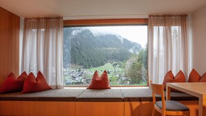 Ein Zimmer mit großem Fenster, Holzdielenboden, einer Bank mit Kissen und einem Tisch mit Stühlen.