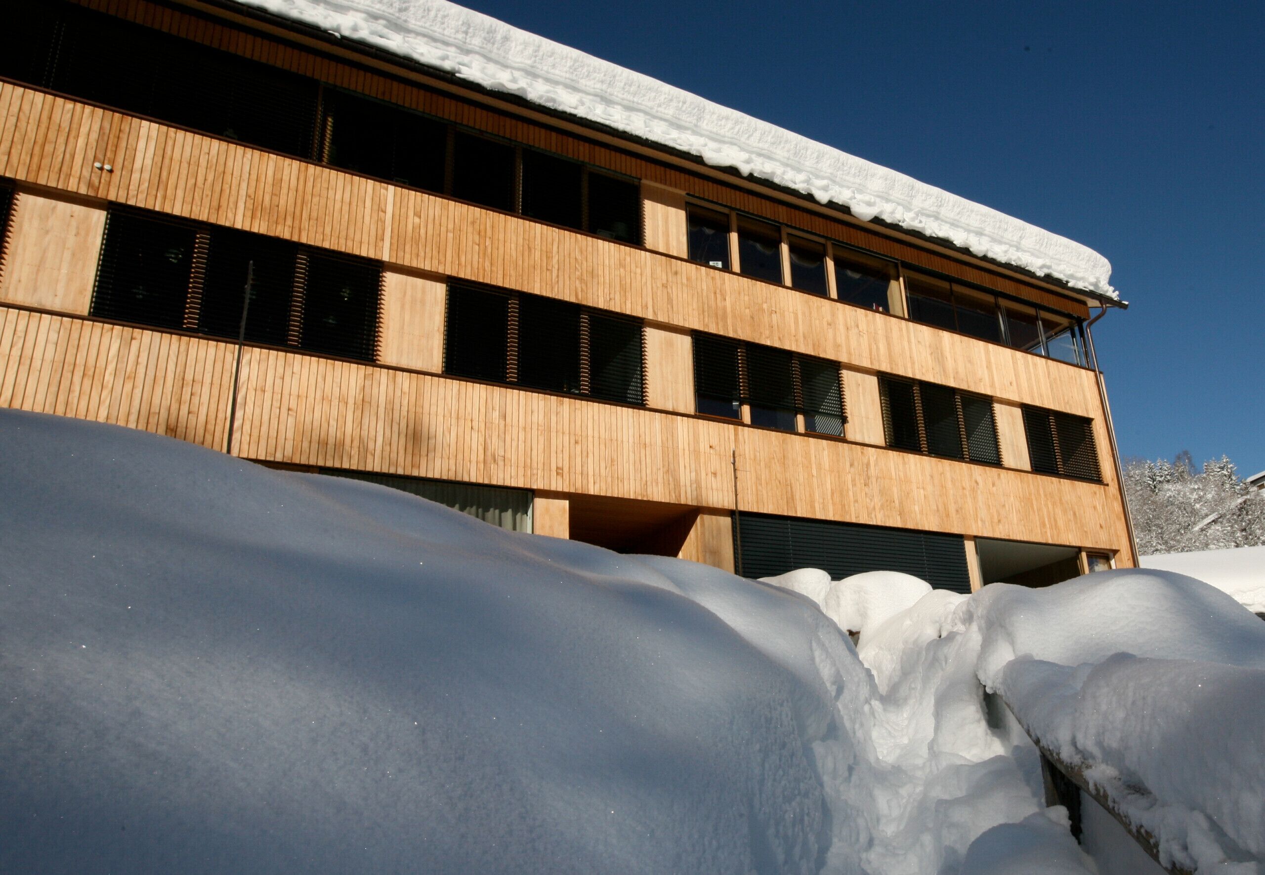 A modern building with a wooden facade and multiple windows, partially covered by snow.