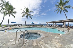 A pool area with lounge chairs and umbrellas, palm trees, and a hot tub.