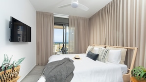 A modern hotel room with a large bed, a ceiling fan, a TV mounted on the wall, and a view of the city through the window. - AmazingApartment W Views of Achziv Beach (Nahariya)