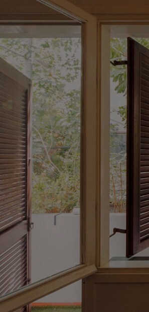 Blick durch ein Fenster mit Holzklappen, der einen Garten mit Bäumen und einer weißen Mauer zeigt.