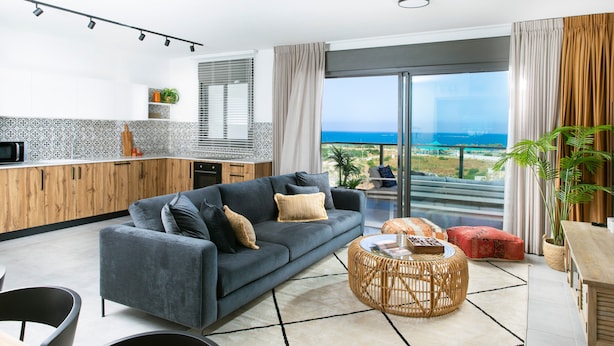 A modern living room with a grey sofa, a round wicker coffee table, and a view of the ocean through large windows. - AmazingApartment W Views of Achziv Beach (Nahariya)