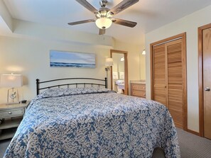 Une chambre à coucher avec un lit, un ventilateur de plafond, une table de chevet, une lampe, un tableau, un miroir, une armoire en bois et une salle de bain visible à travers une porte ouverte.
