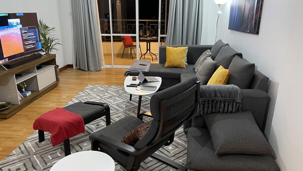 A modern living room with a grey sofa, a small white table, and a red ottoman. There is a TV on a wooden stand, a plant, and a view of the outside through a large window. - perfect apartment (Nairobi)