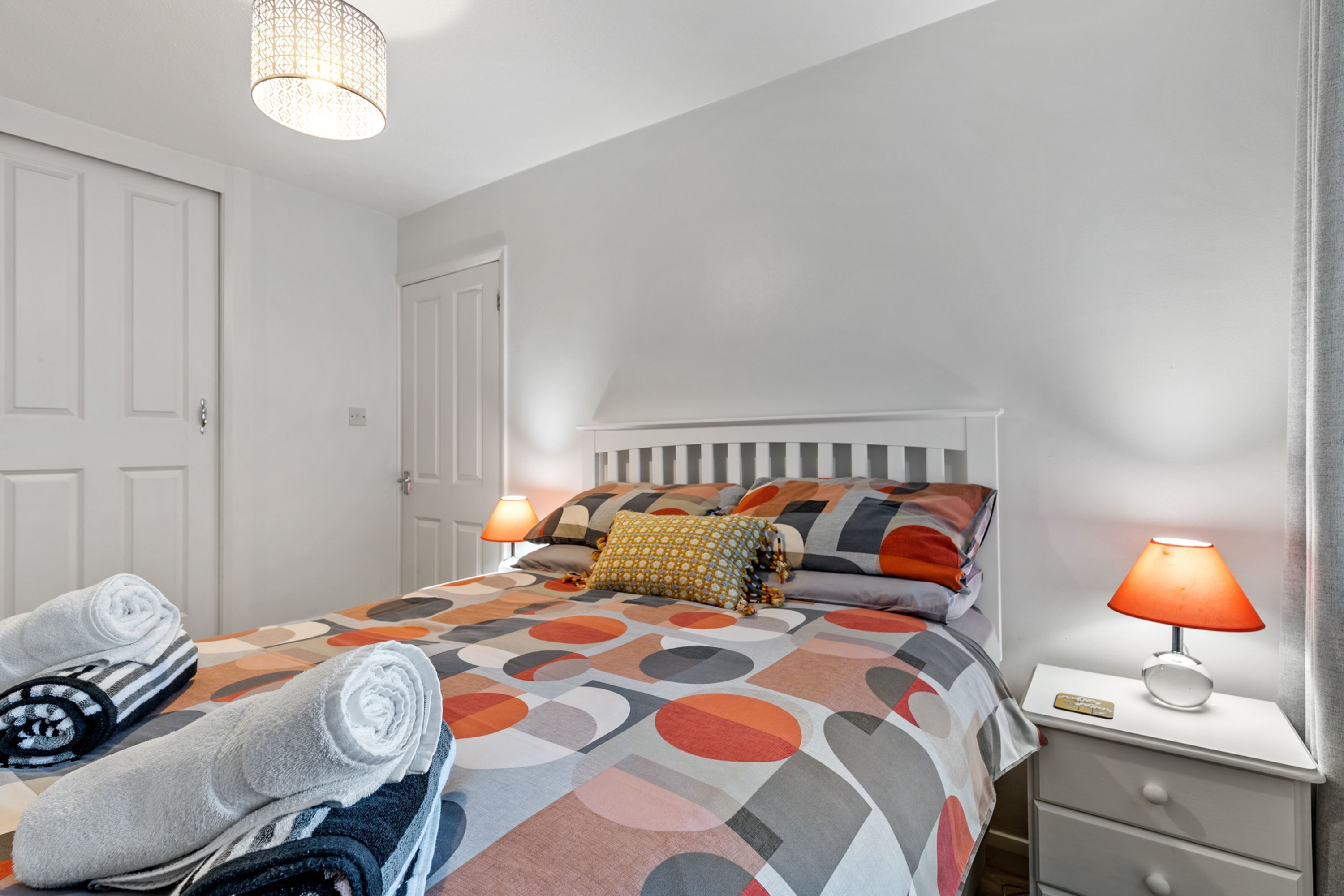 A neatly made bed with a patterned bedspread, two bedside lamps, a nightstand with a lamp, and rolled towels on the floor.