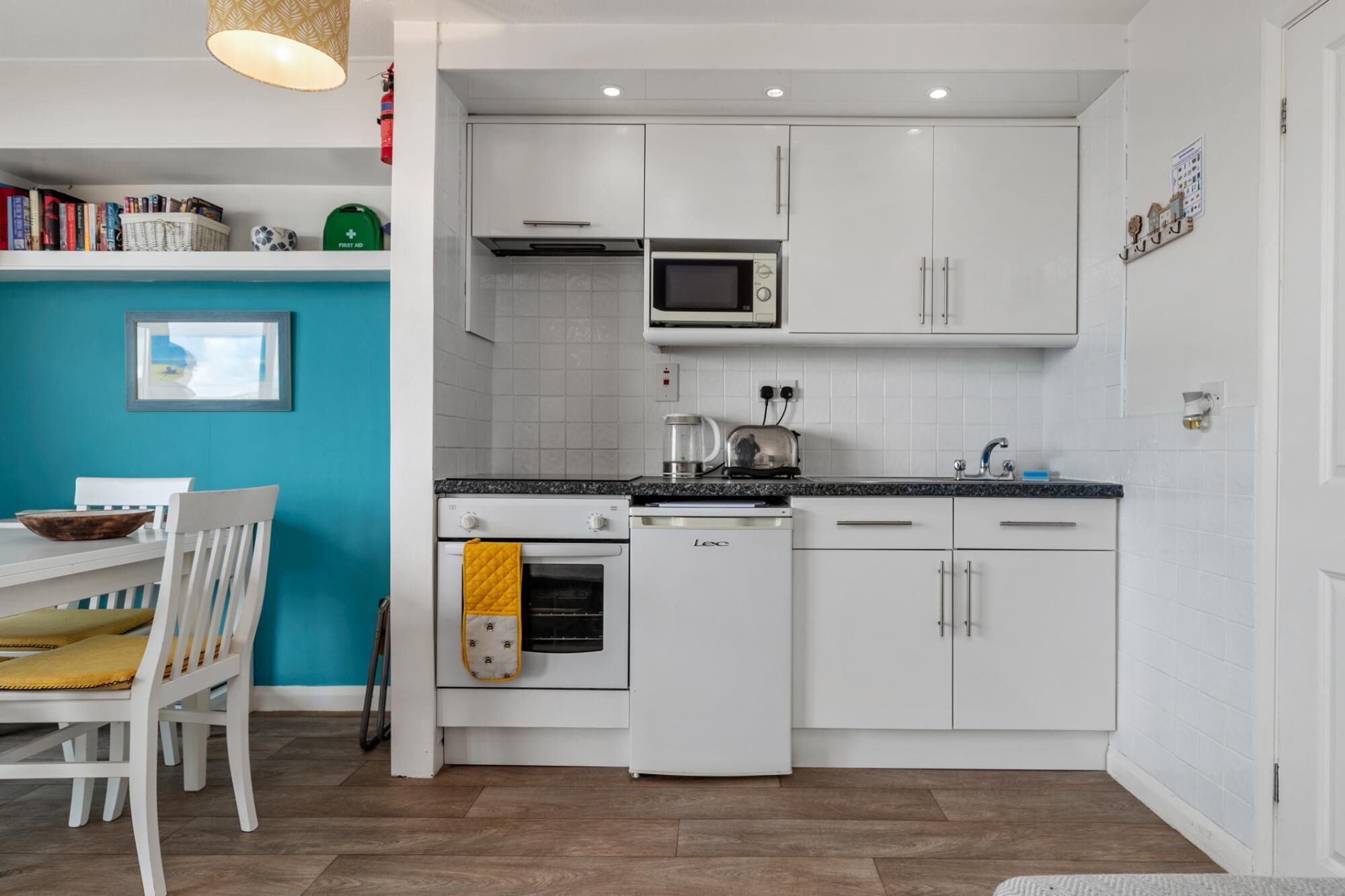 A modern kitchen with white cabinets, a microwave, a kettle, and a dishwasher. There is a dining table with chairs and a blue wall in the background.