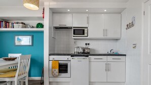 Eine moderne Küche mit weißen Schränken, einer Mikrowelle, einem Wasserkocher und einem Geschirrspüler. Im Hintergrund eine blaue Wand und ein Esstisch mit Stühlen.