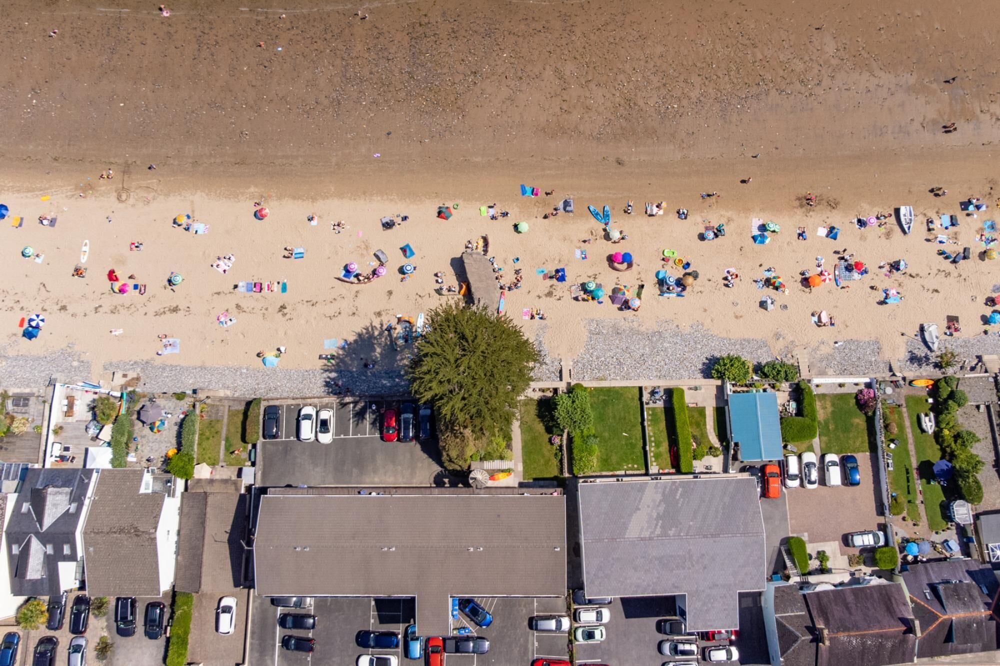 An aerial view of a beach with numerous umbrellas and people. There are several buildings with parking areas in the foreground.
