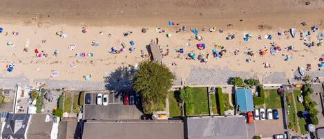 An aerial view of a beach with numerous umbrellas and people. There are several buildings with parking areas in the foreground.
