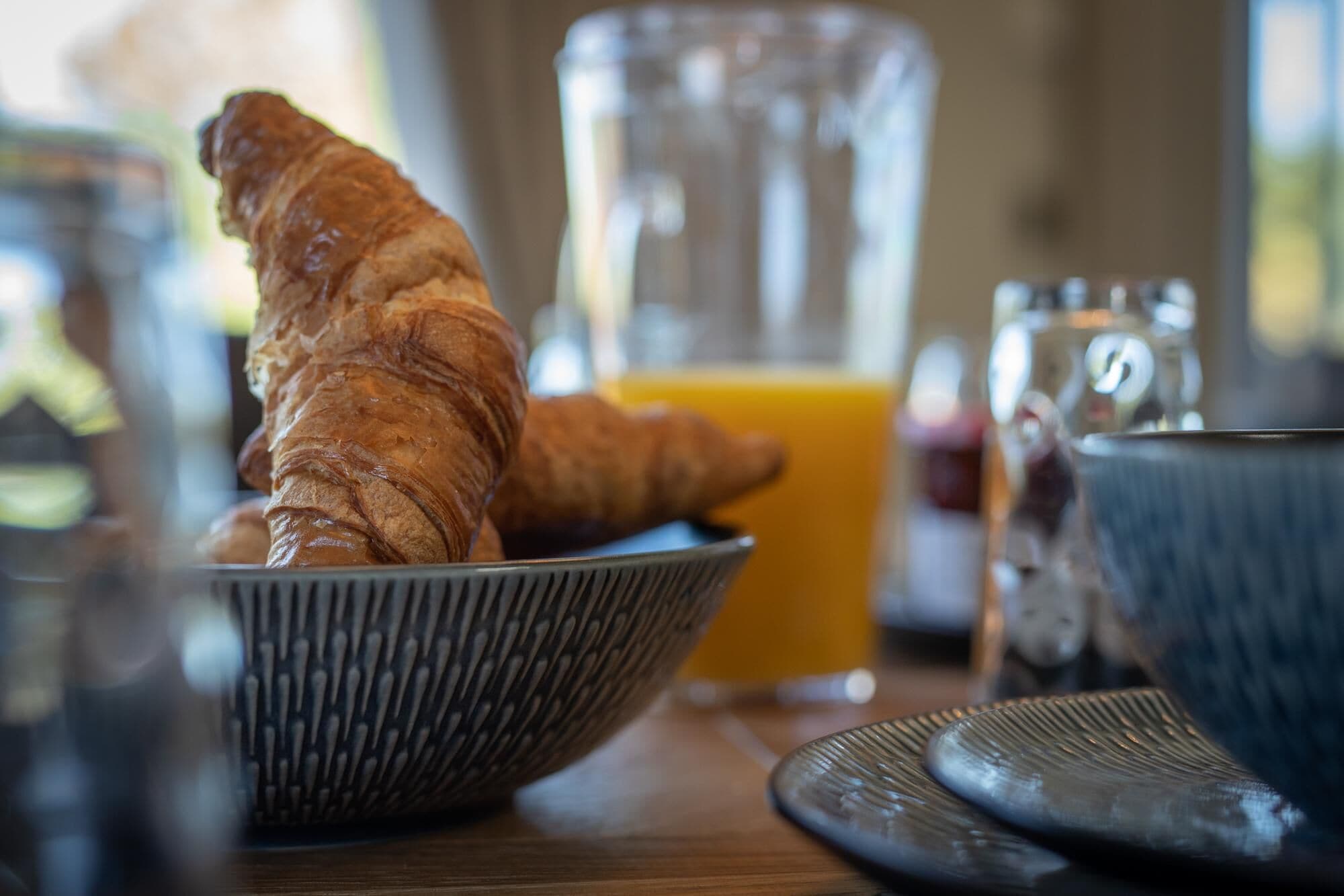 Des croissants dans un bol, un verre de jus d’orange et une tasse en céramique sur une table.