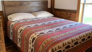 A wooden bed with a patterned quilt and two pillows in a room with wooden flooring and a window. - Post and beam cabin with queen size  and bunk (Shirley Mills)