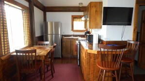 A kitchen with wooden cabinets, a stainless steel refrigerator, a wooden dining table with chairs, and a TV mounted on the wall. - Post and beam cabin with queen size  and bunk (Shirley Mills)