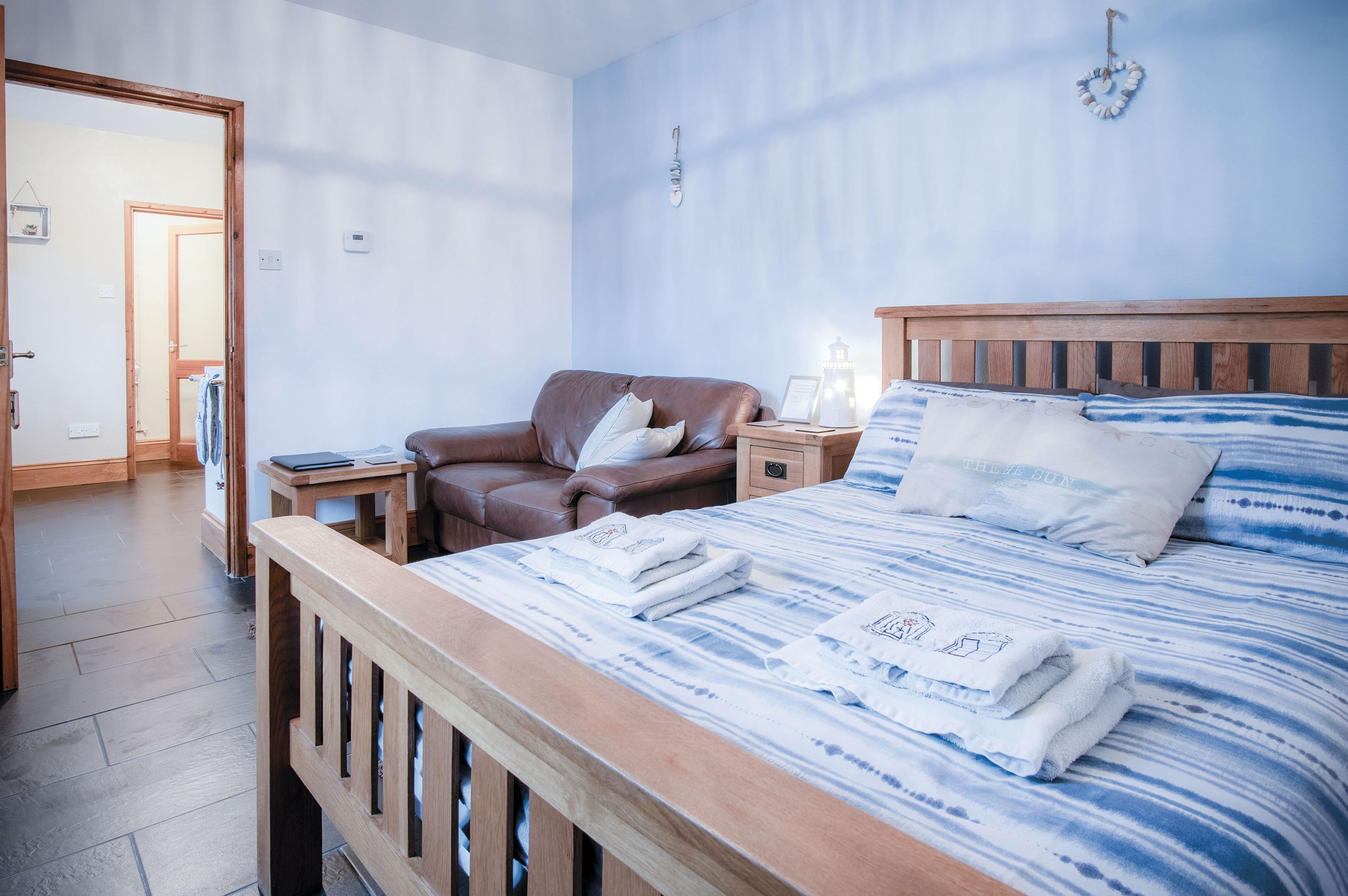 Un dormitorio con una cama de madera, un sillón de cuero marrón, una mesita de noche con una lámpara y una puerta que da a otra habitación.