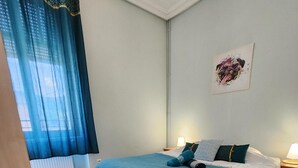 Ein Schlafzimmer mit Bett, Holzboden, einem Fenster mit blauen Vorhängen und einem an der Wand hängenden Kunstwerk.