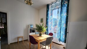 Ein kleiner, heller Raum mit einem Holztisch, zwei Stühlen, einer Topfpflanze, einem weißen Kühlschrank und einem Fenster mit blauen Vorhängen.