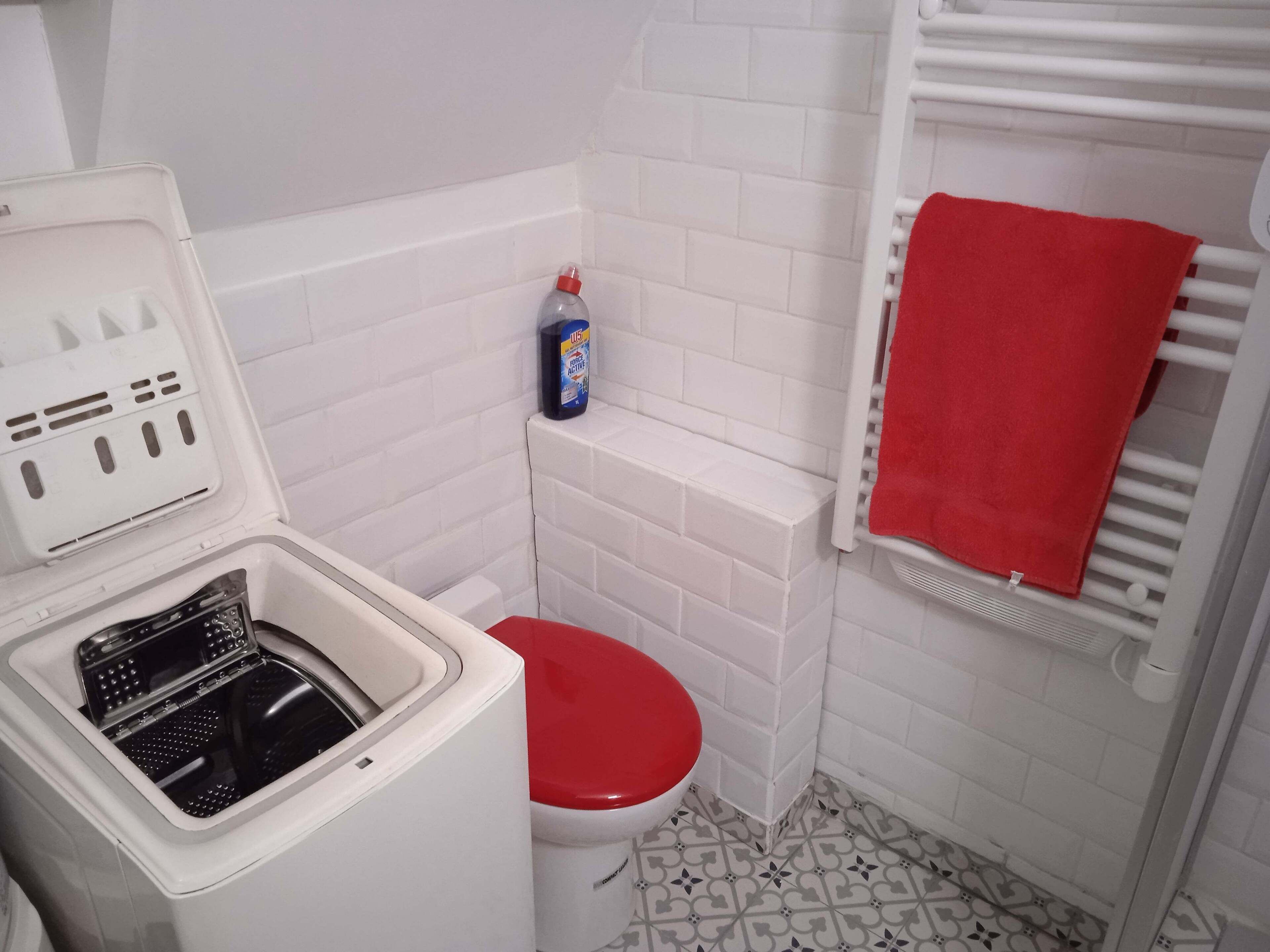 Een kleine badkamer met een wit toilet, een rode zitting, een wasmachine en een fles vloeibare zeep op een plankje.