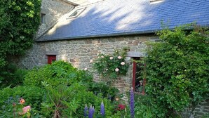 Edifici de pedra amb teulada inclinada, envoltat de vegetació exuberanta i plantes amb flors.