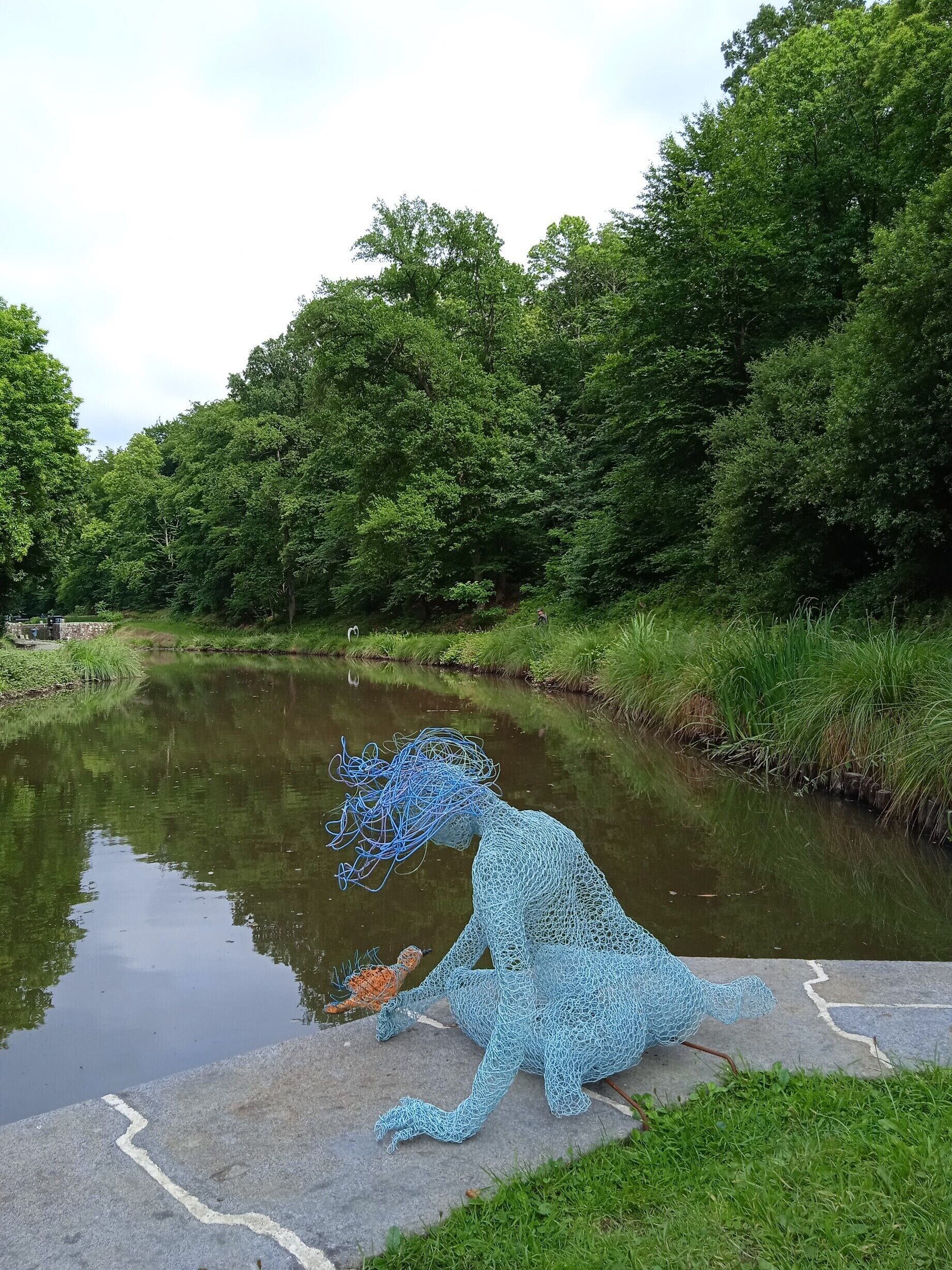 Een sculptuur van een wezen, gemaakt van blauw net, aan een rustige rivier, omgeven door weelderige begroeiing.