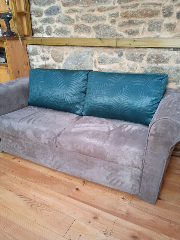 A grey sofa with teal cushions against a stone wall, wooden floor, and a wooden shelf with a drill. - Le Rocher Cordier (Bonnemain)