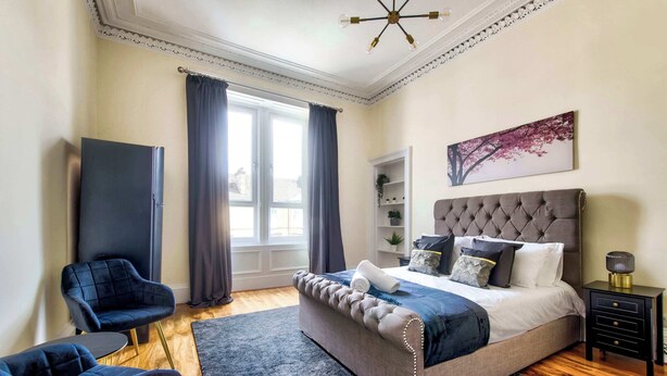Een slaapkamer met een bed, een stoel, een nachtkastje, een raam met gordijnen en een aan de muur hangend schilderij.