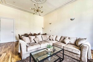 A living room with a sectional sofa, a glass coffee table, a chandelier, and a wooden floor.