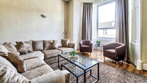 Een woonkamer met een hoekbank, een glazen salontafel, een bijzettafeltje en twee fauteuils.