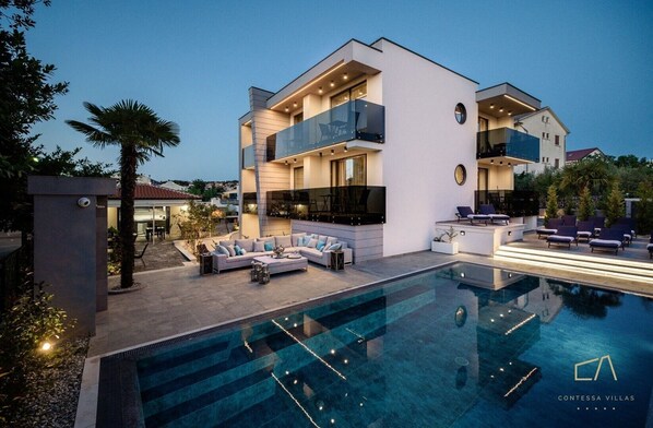 Eine moderne Villa mit Schwimmbad, Sitzgelegenheiten im Freien und einem gut beleuchteten Balkon.