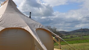 Een grote, beige tent met een puntige kap, midden in een grasveld met een heuvelachtig landschap op de achtergrond.