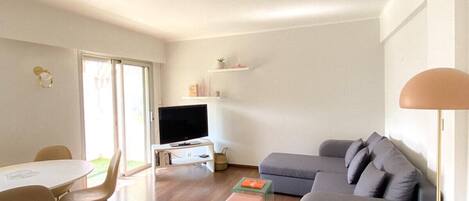 A modern living room with a grey sofa, a round dining table with chairs, a TV on a white stand, and a floor lamp.