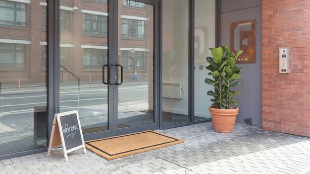 A modern building entrance with glass doors, a welcome sign, a potted plant, and a brick wall. - Modern 1BED Central Birmingham Apartment- Sleeps 4 (Birmingham)