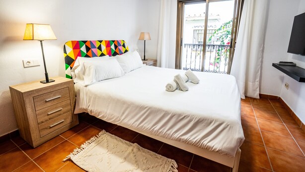 A neatly made bed with white linens, a colorful geometric headboard, and a pair of rolled towels on the bed. There is a wooden nightstand with a lamp and a drawer, and a white rug on the floor. - Marbella Village | Studio with balcony (Marbella)
