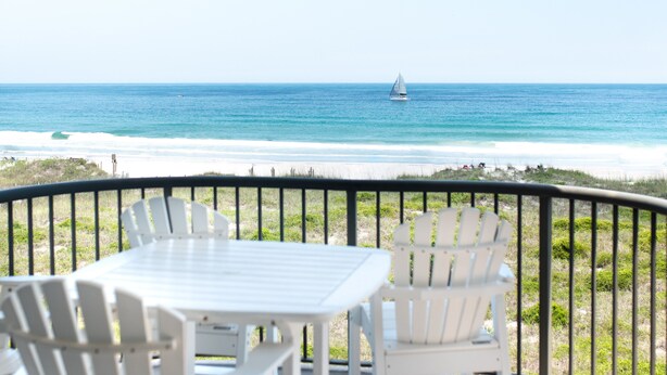 A deck with white chairs and a table overlooking a beach with a sailboat in the distance. - 3 Bed~3 Bath~Sleeps 8~Duneridge Condo~Wrightsville Beach, NC (Wrightsville Beach)