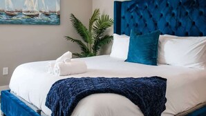 Ein ordentlich bezogenes Bett mit weißen Laken und einem blauen Kopfteil, ein Segelbootgemälde an der Wand und ein Blumentopf neben dem Bett.