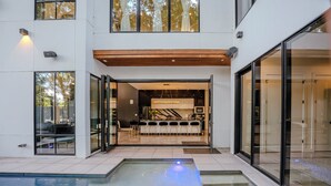 Un bâtiment moderne doté d’une piscine, de grandes fenêtres et d’un espace cuisine visible à travers les portes ouvertes.