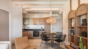 A modern kitchen with wooden cabinets, a dining area with a round table and chairs, a wicker chair, and a bookshelf with decorative items. - Sea View Villa w/Private Pool, 400m to the beach (Heraklion, Crete)