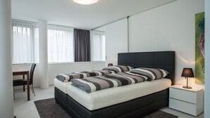A modern bedroom with a black bed, a bedside table with a lamp, a desk with a chair, and a large window with curtains.