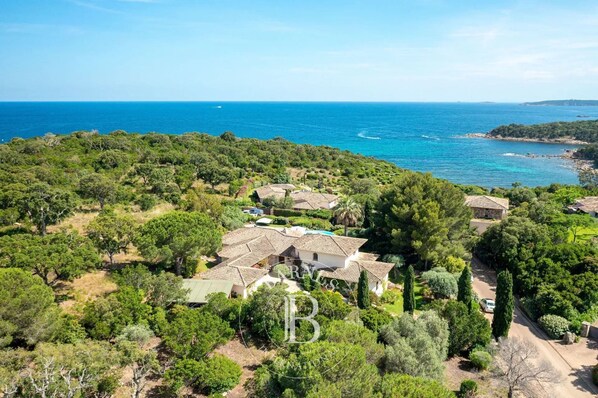 A coastal property with multiple buildings surrounded by dense greenery and a clear view of the sea.