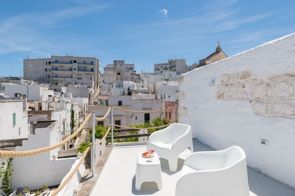 A rooftop terrace with white chairs and a small table, overlooking a densely packed urban area with buildings and a church. - Corso Italia Deluxe Apartment by Wonderful Italy (Ostuni)