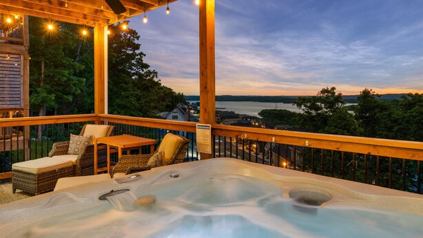 En træterrasse med jacuzzi, udendørs møbler og udsigt over en sø i skumringen.