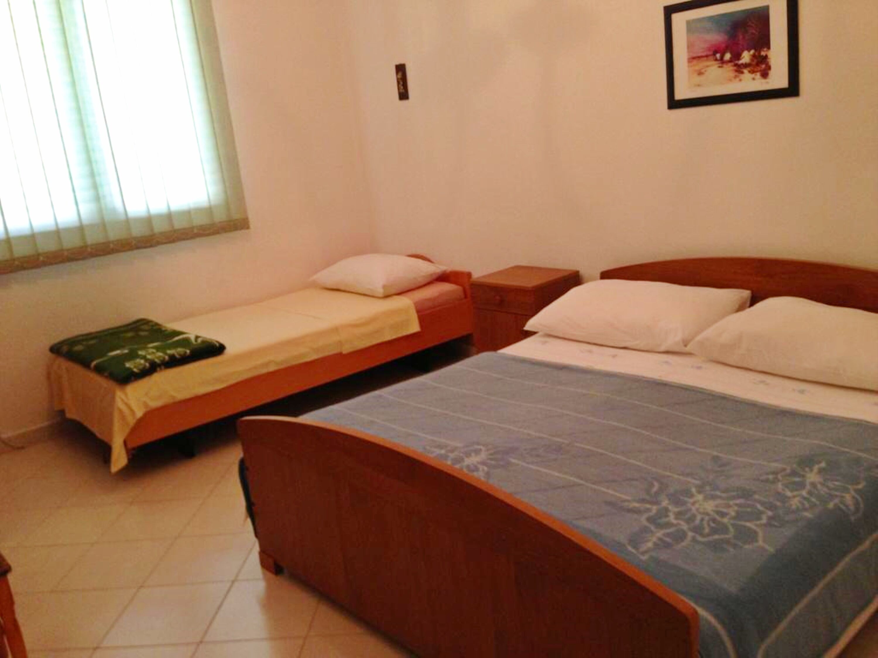 A bedroom with two beds, a window with blinds, a small bedside table, and a framed picture on the wall.