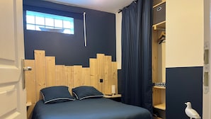 A bedroom with a bed, wooden headboard, a window with blinds, a nightstand, a wardrobe, and a small white bird sculpture. - Bélicine, seafront apartment for 5 people  (LE CROTOY)