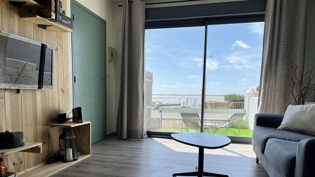 A modern living room with a large window, a grey sofa, a small round table, and wooden shelving. - Bélicine, seafront apartment for 5 people  (LE CROTOY)