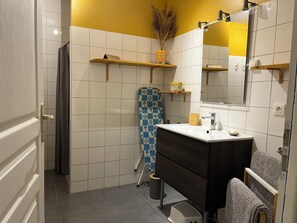 A bathroom with white and yellow tiled walls, a wooden vanity, a mirror, and a shower area. - Bélicine, Ocean View Apartment for 5 People (LE CROTOY)