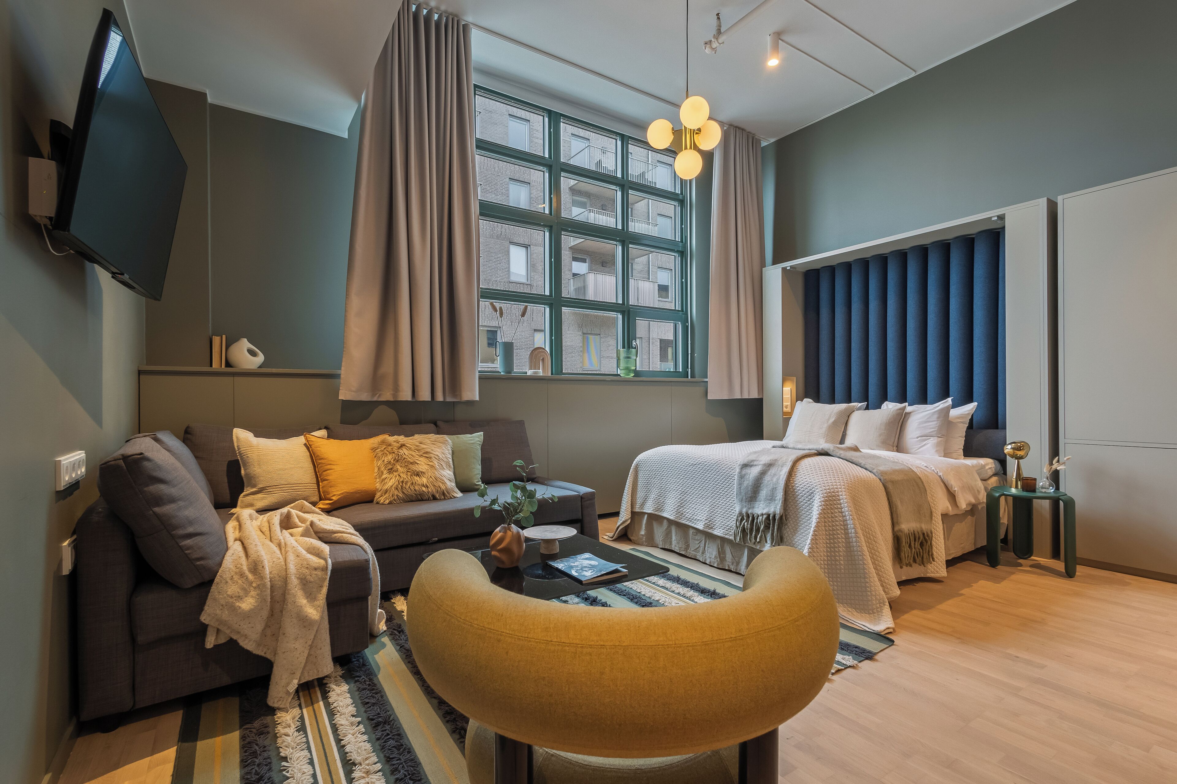 Een moderne hotelkamer met een bank, een bed, een televisie en een groot raam met gordijnen.