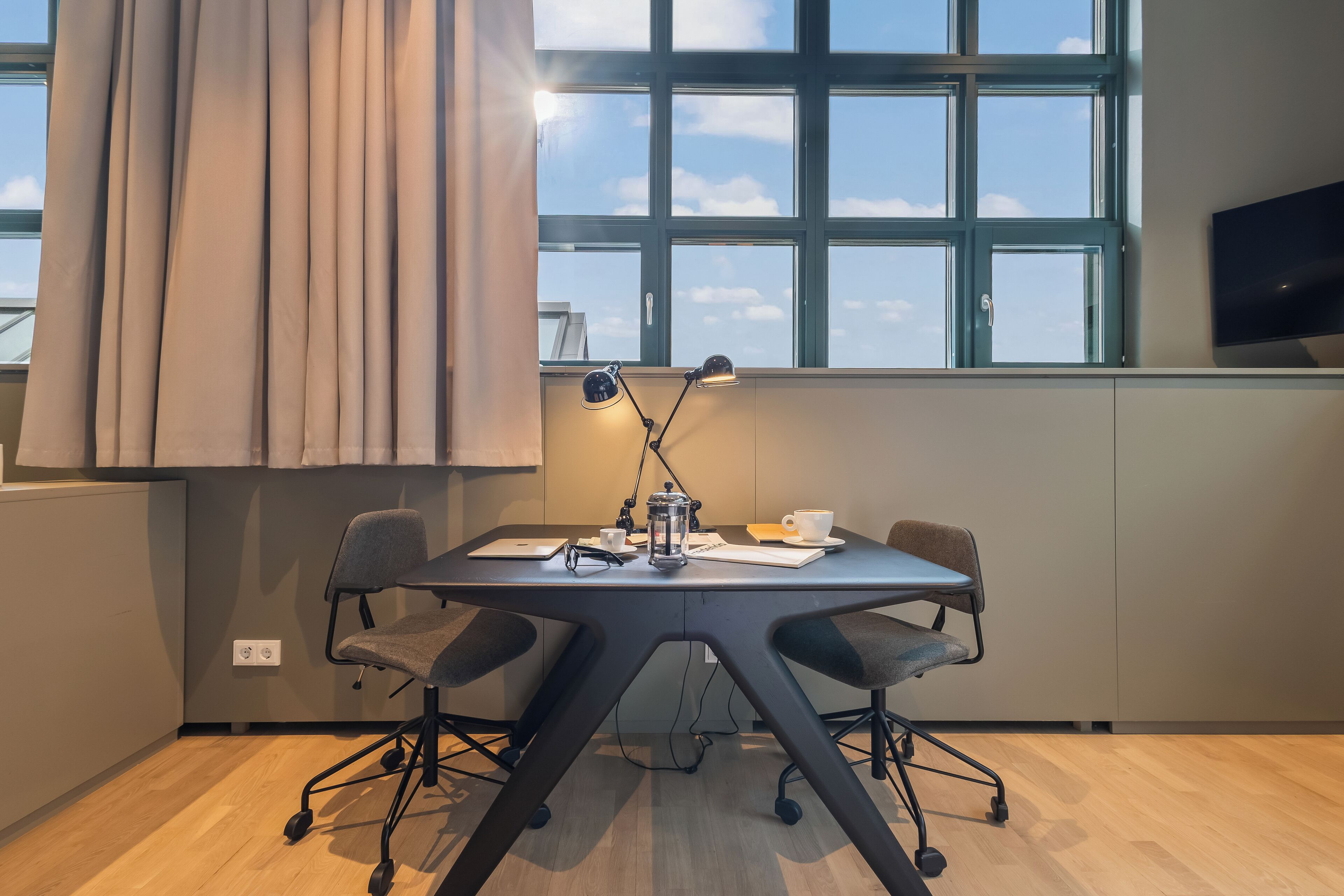 A modern meeting room with a large window, a table with chairs, a coffee pot, and a laptop.