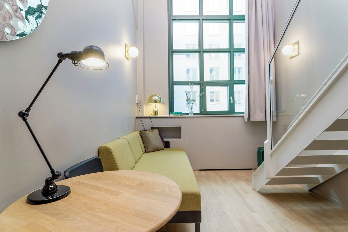 A modern room with a round wooden table, a green sofa, a black desk lamp, a window with curtains, and a staircase.