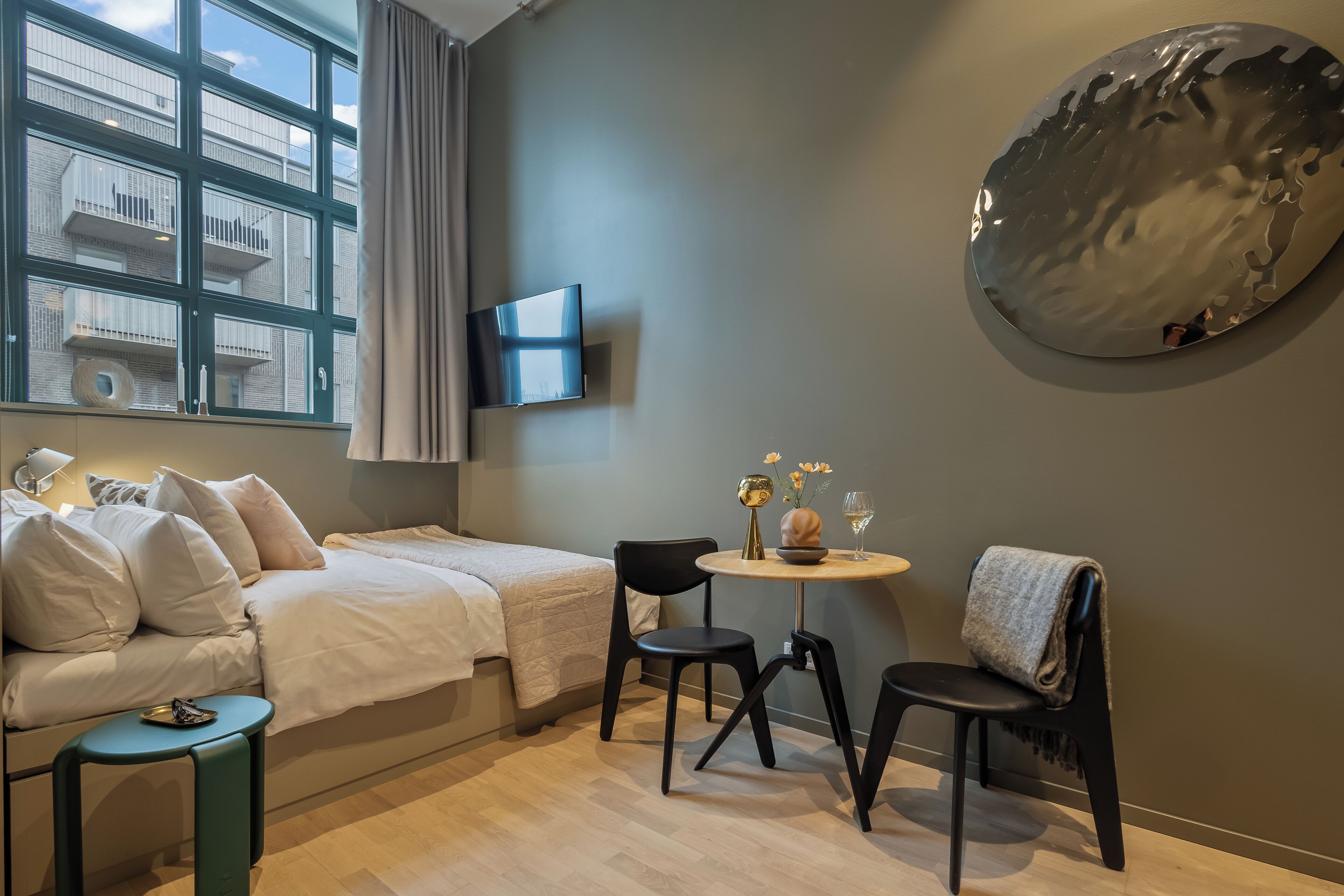 Ein modernes, kompaktes Zimmer mit einem Bett, einem kleinen Tisch mit Stühlen, einem an der Wand montierten Fernseher und einem großen, abstrakten Kunstwerk.