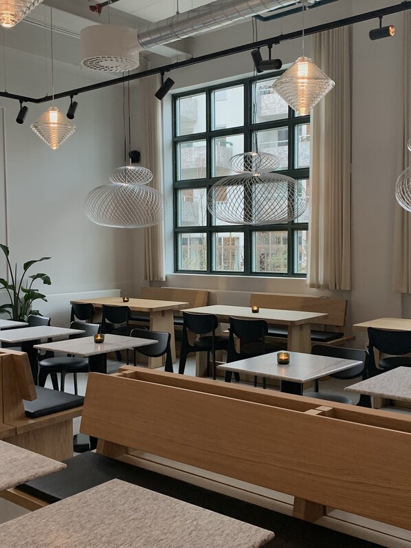 A restaurant interior with multiple tables and benches, hanging pendant lights, and a large window with curtains.